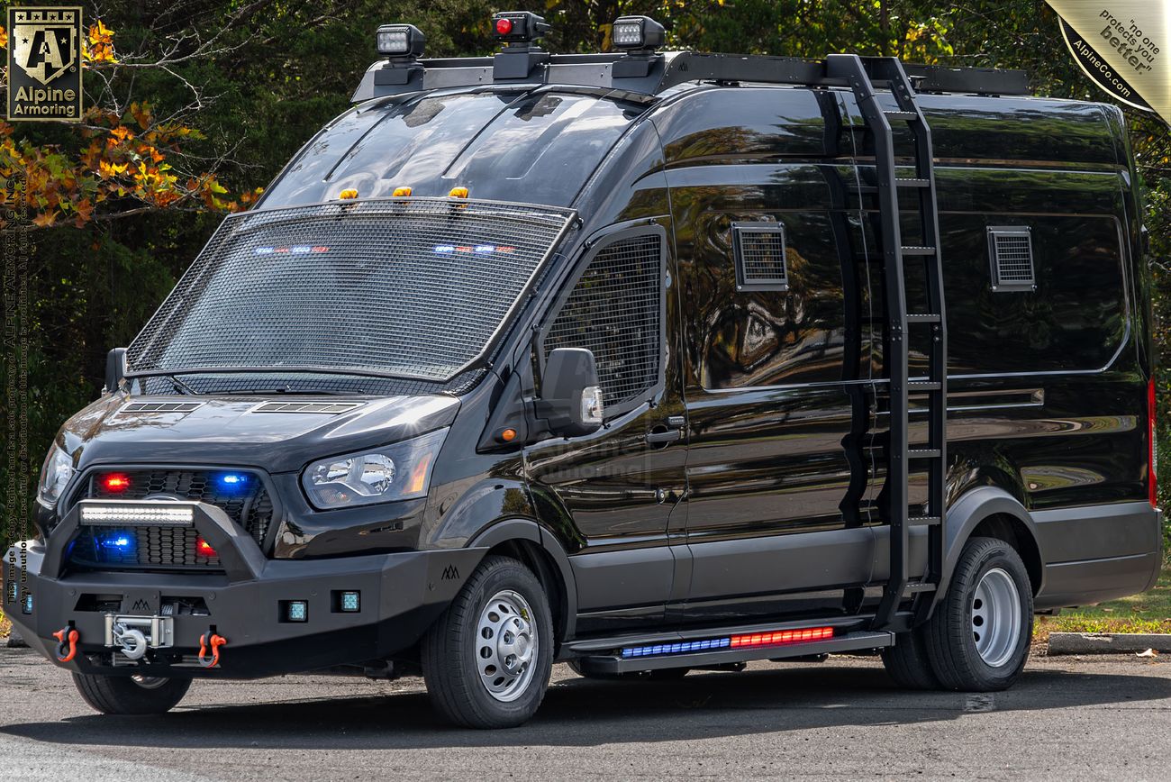 A black, armored Pointer van with red, blue, and white lights on its grille and roof is parked on a road. The vehicle features metal mesh over the windows and additional equipment attached to the front and sides.