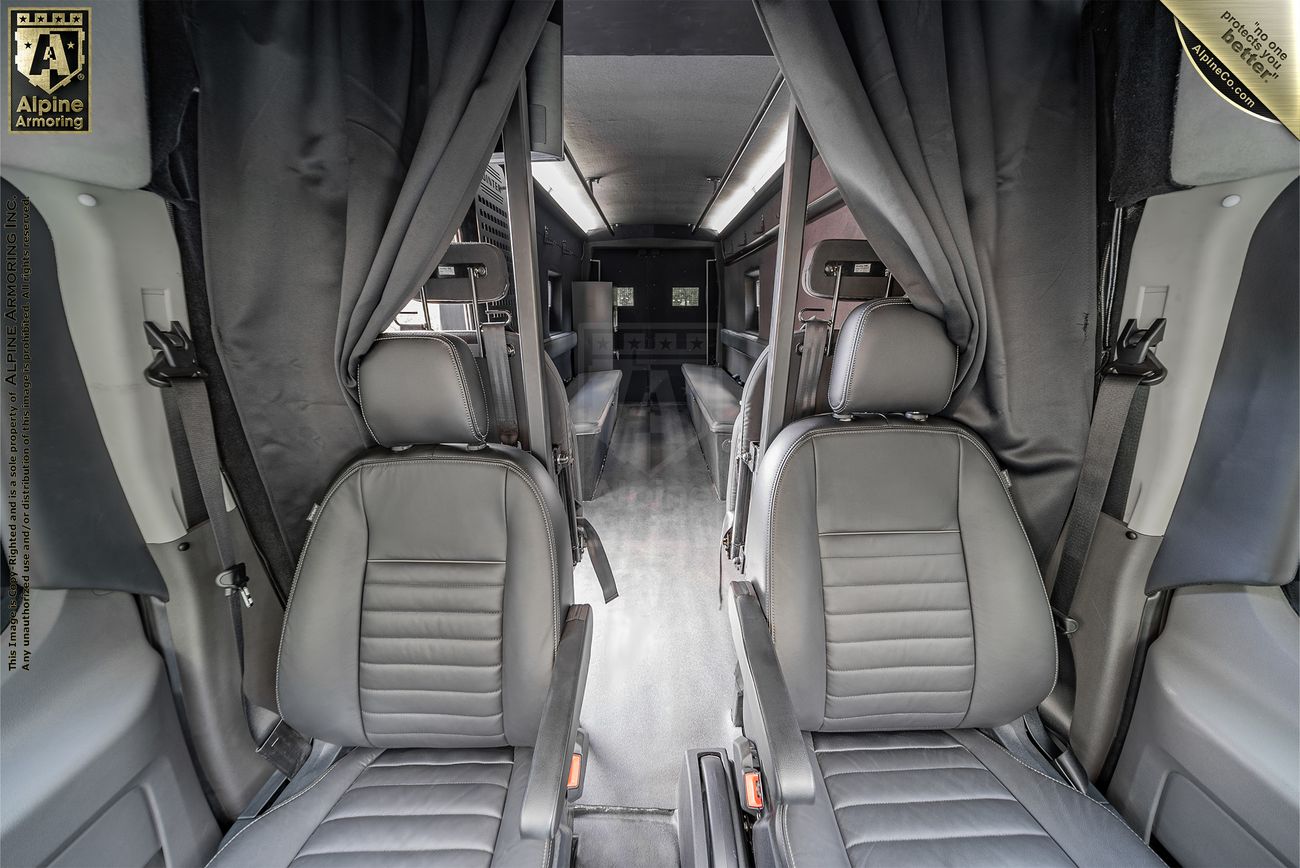 A view of an armored Pointer van featuring two gray leather seats in the foreground with additional seating and a partitioned area visible in the background. Curtains are drawn on either side.