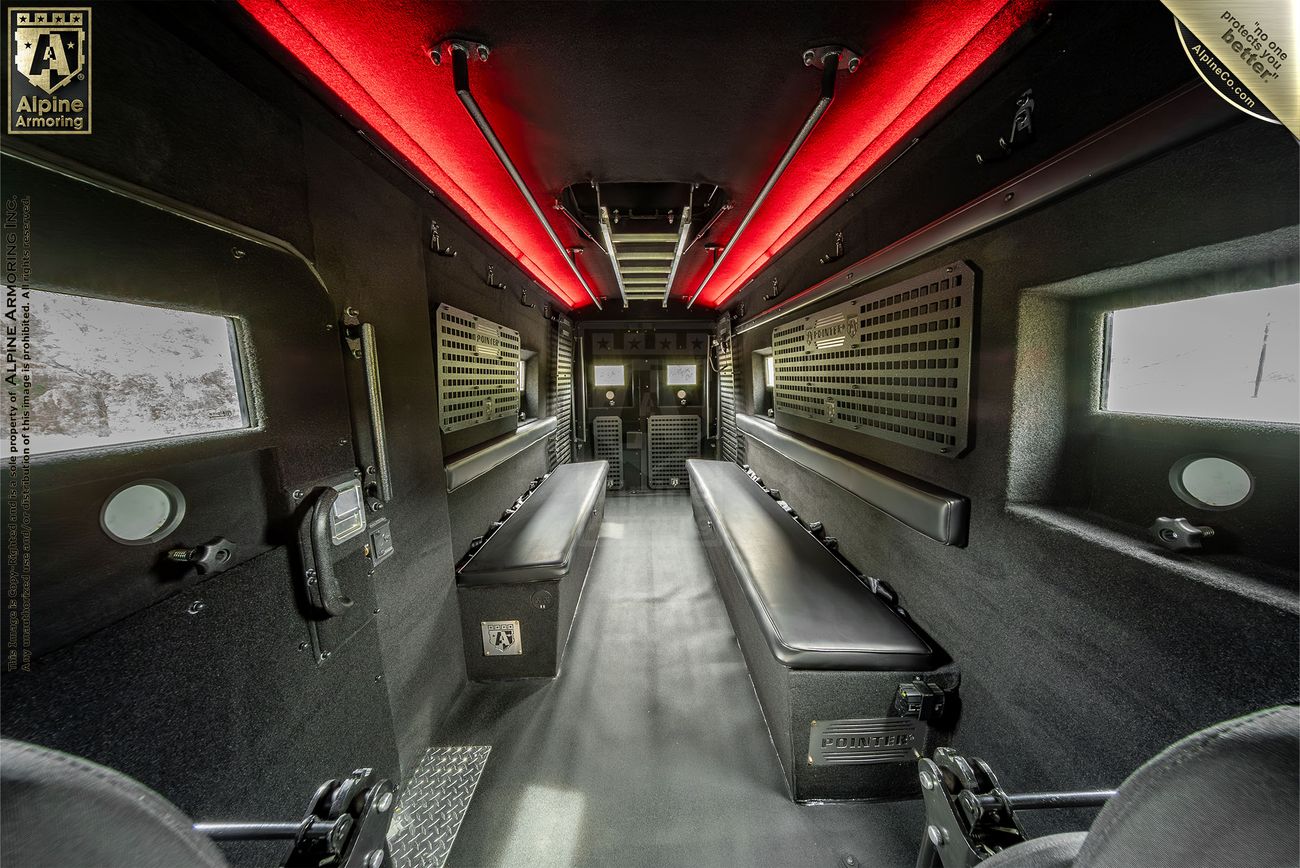 Interior view of an armored Pointer van showing two rows of black bench seating, red accent lighting on the ceiling, and various equipment mounted on the walls.