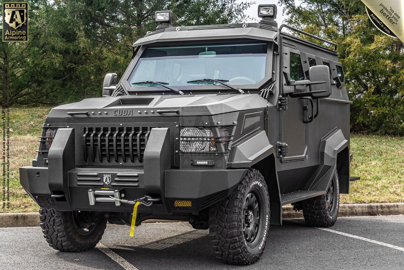 A black armored CUDA SWAT vehicle with rugged tires, angular body panels, and a front grille guard is parked on a paved area with greenery in the background.