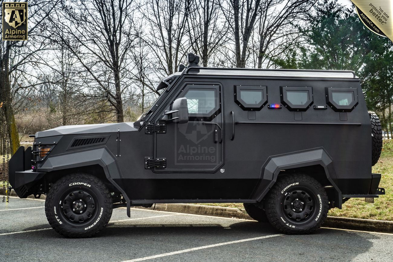 A matte black, armored tactical CUDA is parked outdoors on a paved surface, with trees and grass visible in the background. The vehicle features tinted windows and heavy-duty tires.