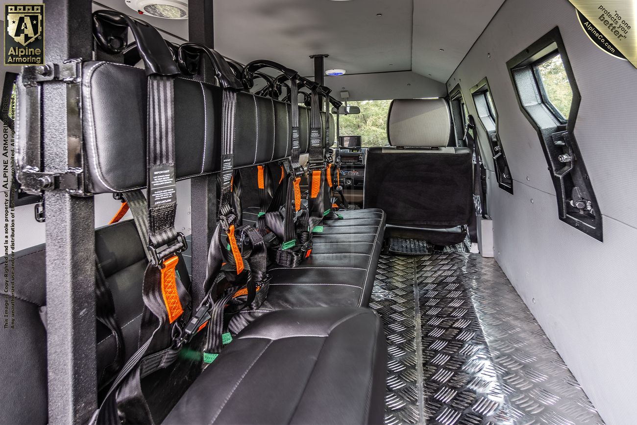 Interior view of an armored CUDA from Alpine Armoring with black padded seats, harnesses, and metal flooring, showing a driver's seat and steering wheel in the front. The vehicle has small windows along the sides.
