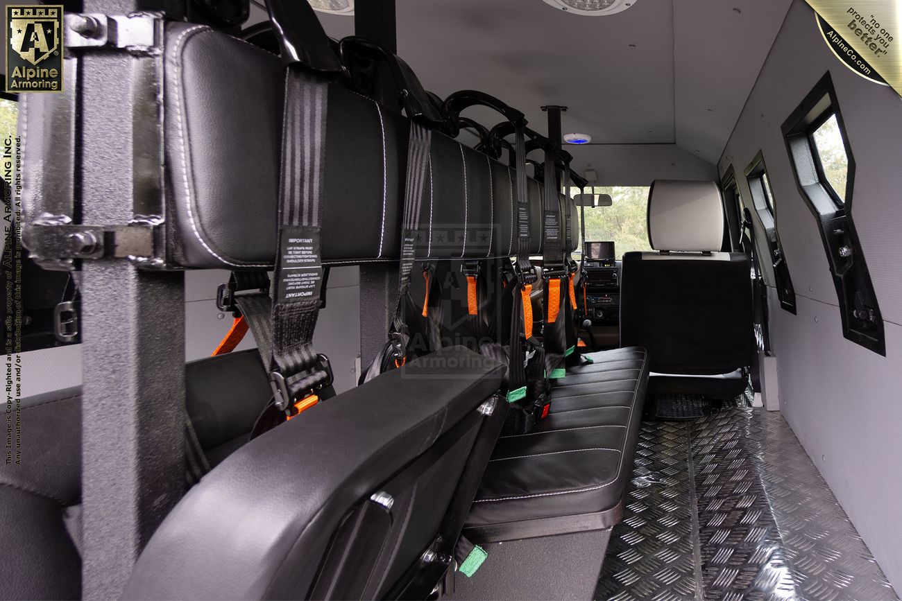 Interior of a CUDA SWAT vehicle with black leather seats and orange seat belts, facing a wall. The floor appears to be metal, and there is a window towards the front.