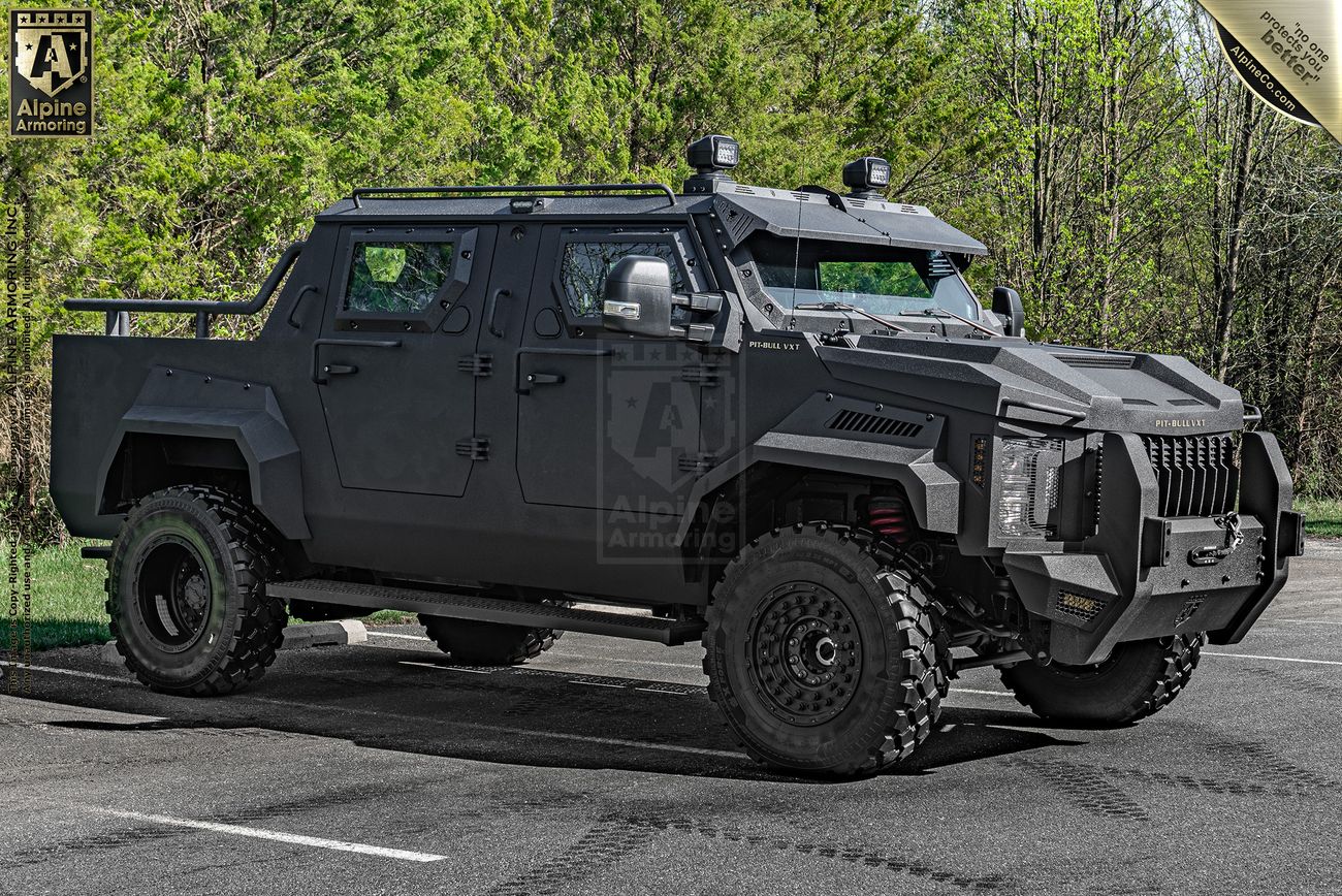 A matte black armored Pit-Bull VXT SWAT truck with rugged tires is parked on a paved road, with green foliage in the background. The vehicle has a robust design and multiple lights on top.