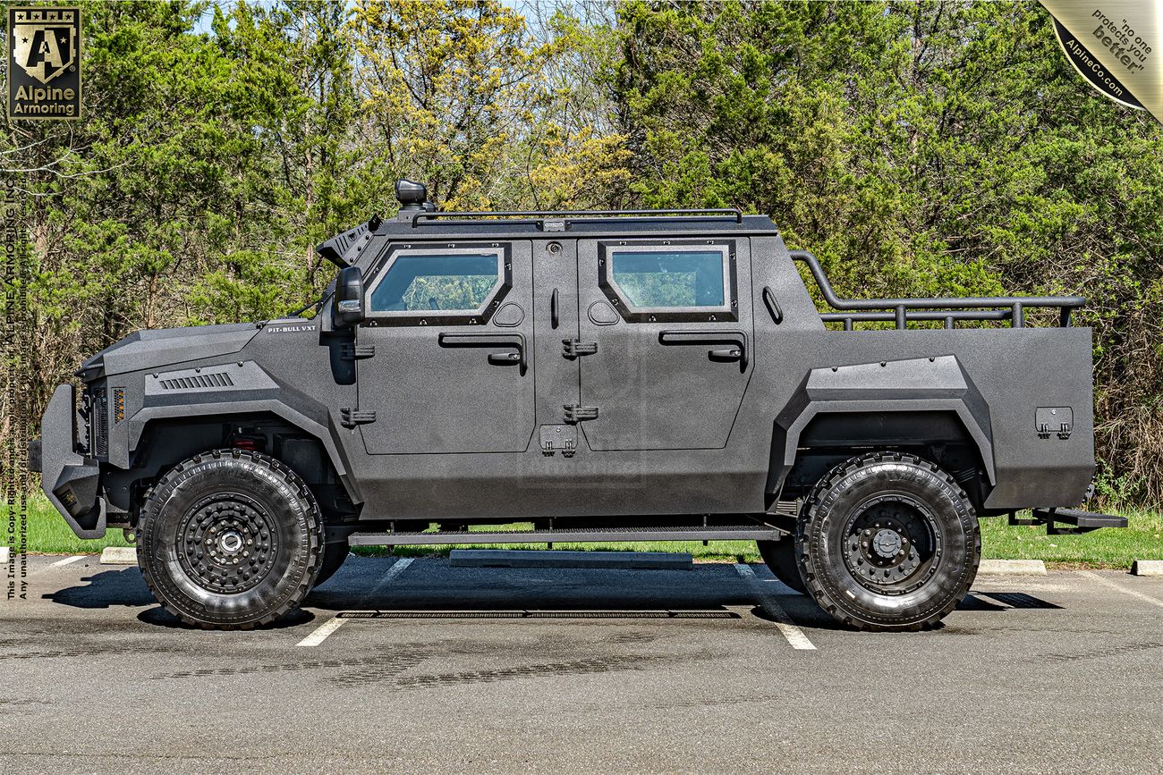 A large armored Pit-Bull VXT with rugged tires is parked in an open area. It has a matte black exterior and reinforced windows, built for heavy-duty use and protection. Trees are visible in the background.