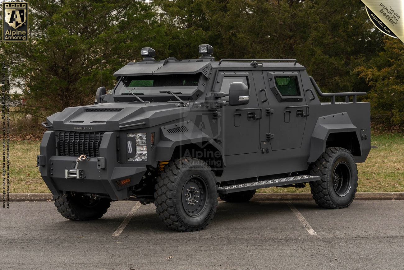 A matte black armored and bulletproof Pit-Bull VXT with rugged tires and reinforced windows is parked on a paved surface, surrounded by greenery. An "Alpine Armouring" logo is visible on the side of the vehicle.