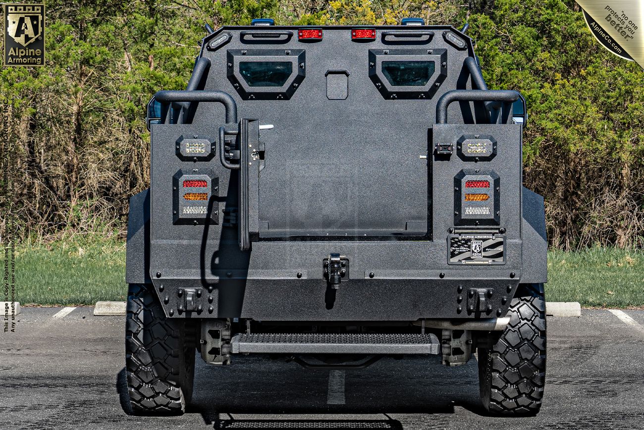 A heavily armored black Pit-Bull VXT SWAT truck is parked, rear view showing thick tires, tail lights, emergency lights on top, and a reinforced door. Trees and asphalt are visible in the background.