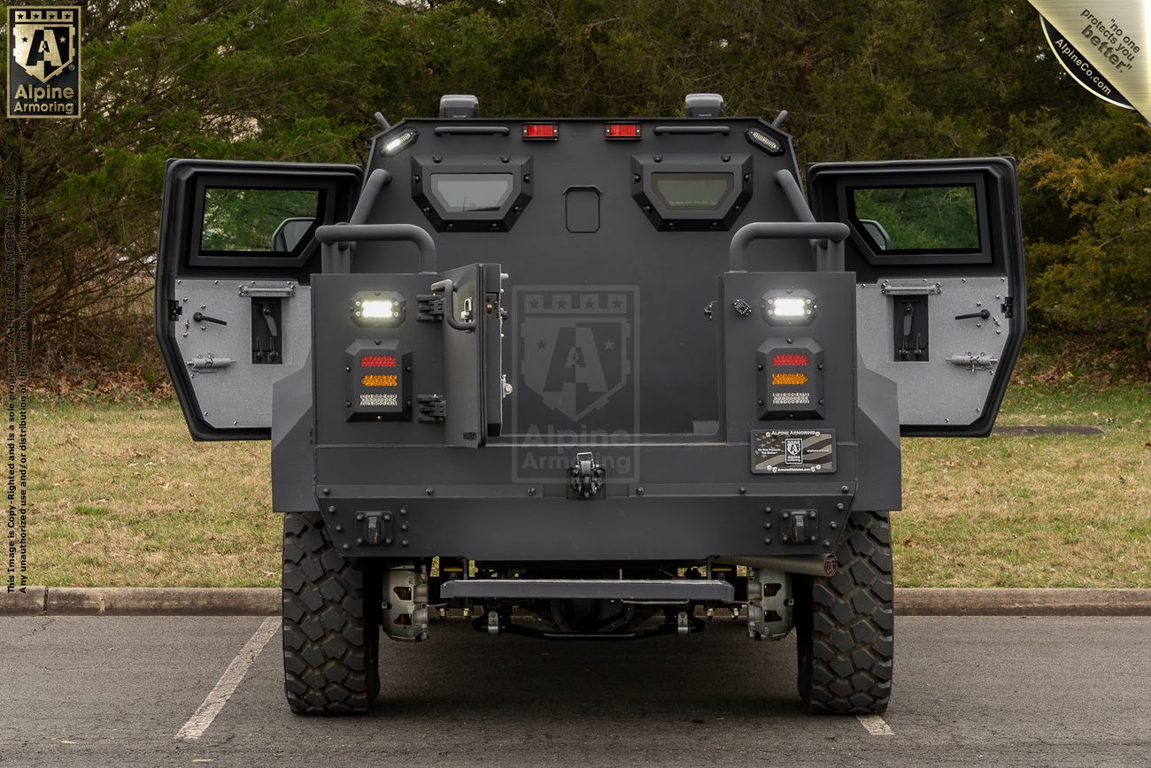 A rear view of a heavily armored black Pit-Bull VXT APC from Alpine Armoring, with its back doors open. The vehicle is parked on a paved area with greenery in the background.