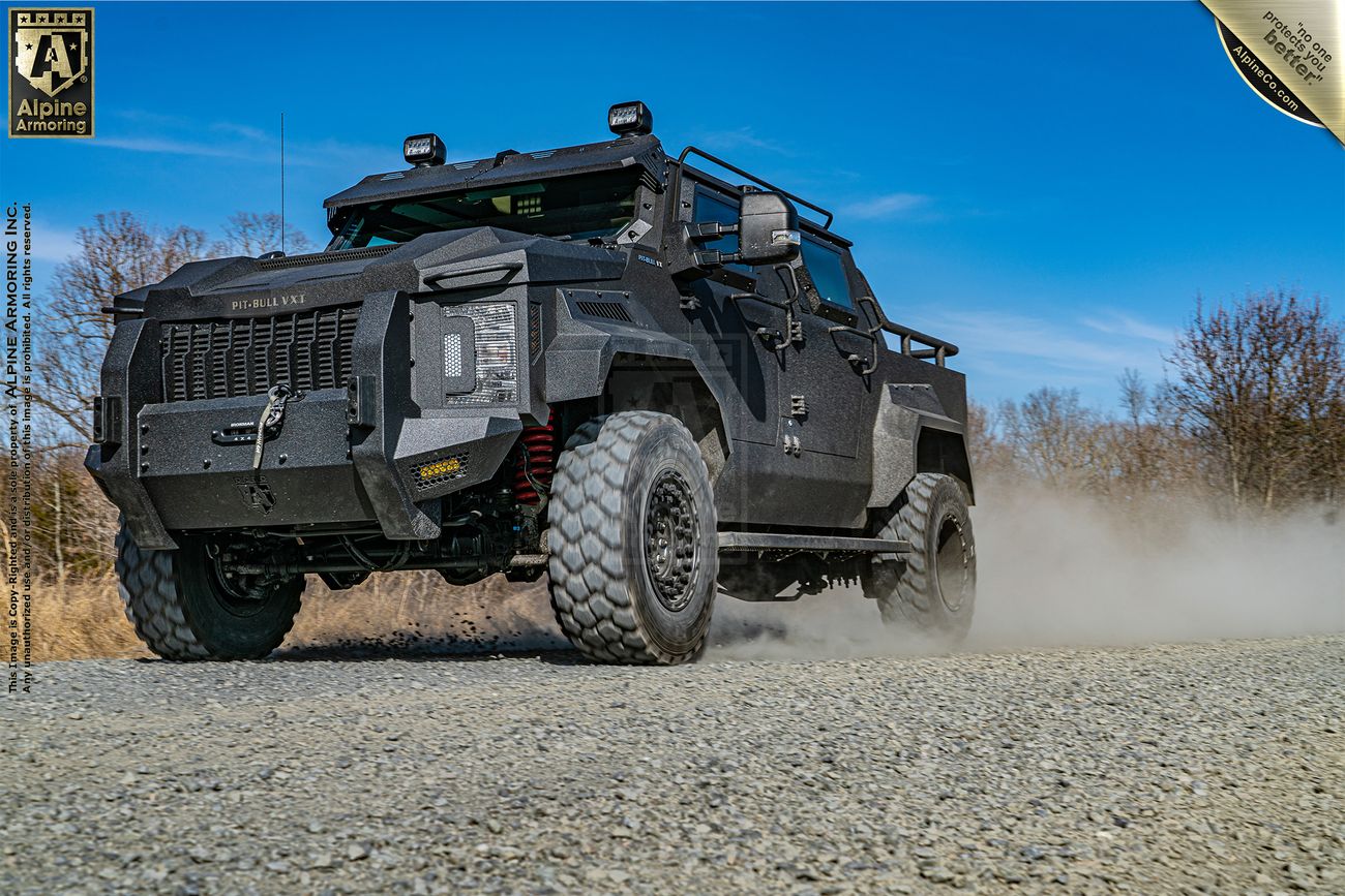 A heavily-armored, all-terrain, bulletproof Pit-Bull VXT drives on a gravel path. The vehicle has large, rugged tires and a black exterior. Sparse trees are visible in the background under a clear blue sky.