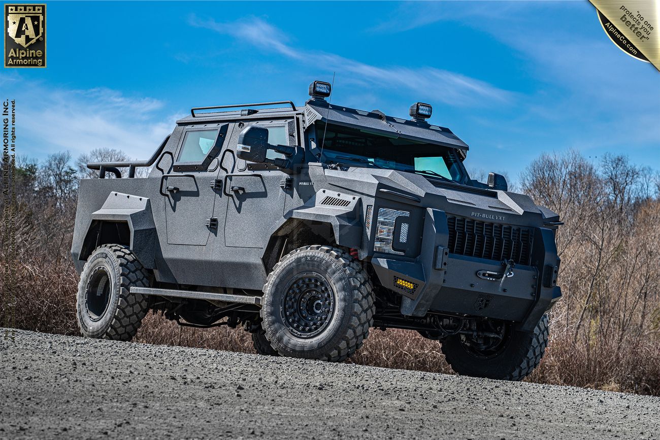 A heavily armored, Pit-Bull VXT vehicle with large off-road tires parked outdoors on a gravel surface with a partially cloudy sky and bare trees in the background.