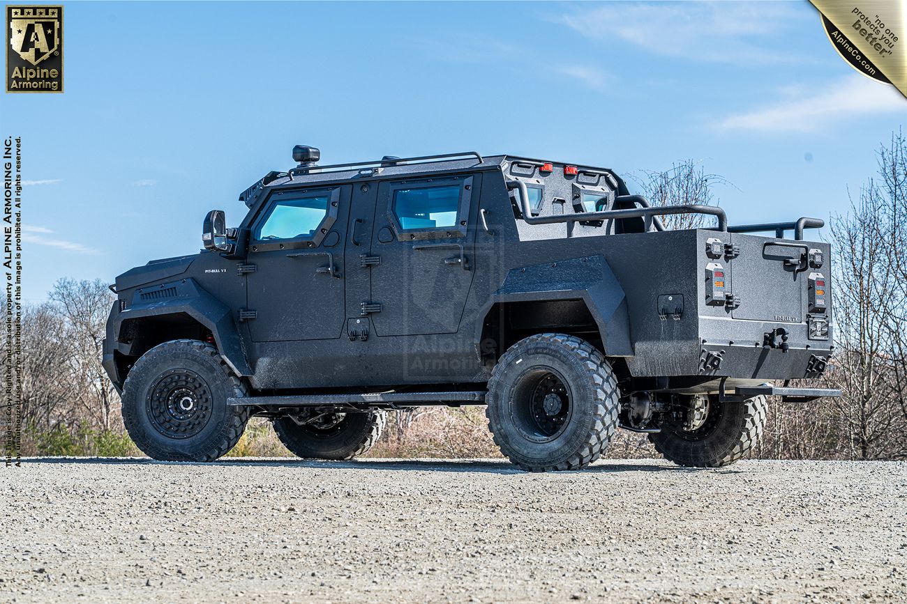 A black armored Pit-Bull VXT, designed by Alpine Armoring, is parked on a gravel surface, with a clear blue sky and sparse vegetation in the background.