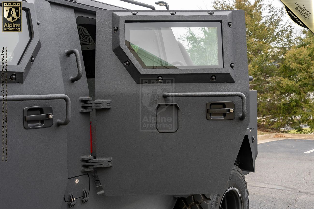 Close-up view of an armored Pit-Bull VXT's side, showing the heavy-duty door with a small window and latch mechanism. Trees are visible in the background.