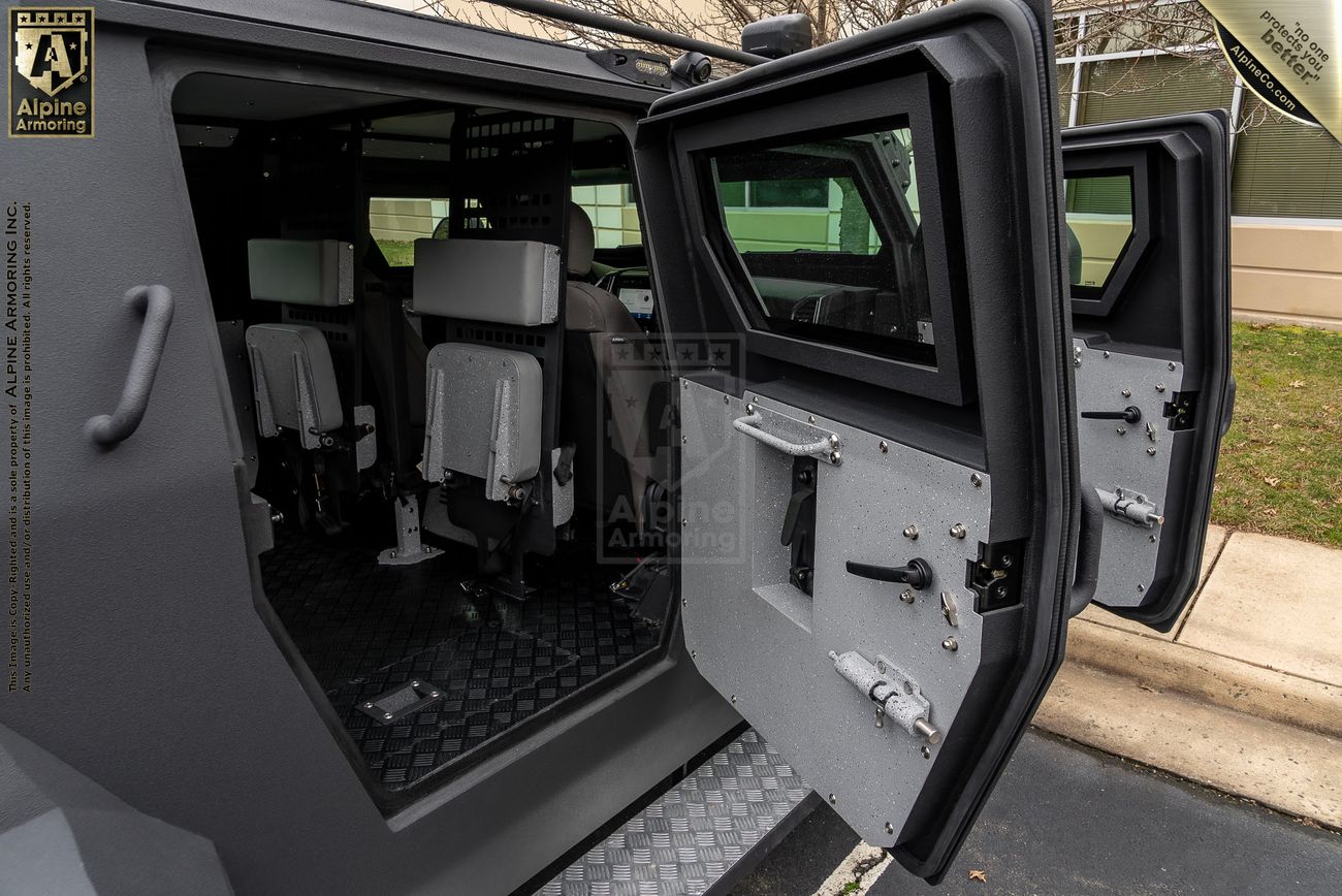 A view of an armored Pit-Bull VXT with its doors open, showing the interior seats and protective design features. The vehicle is parked outdoors on a paved surface beside a building and a grassy area.