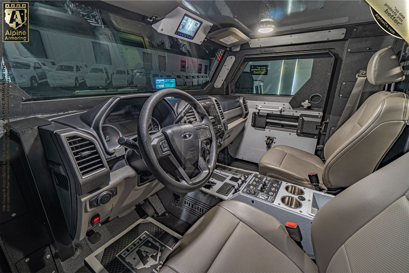 Interior of a Pit-Bull VXT SWAT vehicle, featuring a steering wheel, control panels, rear-view screen, and beige leather seats. The vehicle's robust structural reinforcements are visible.