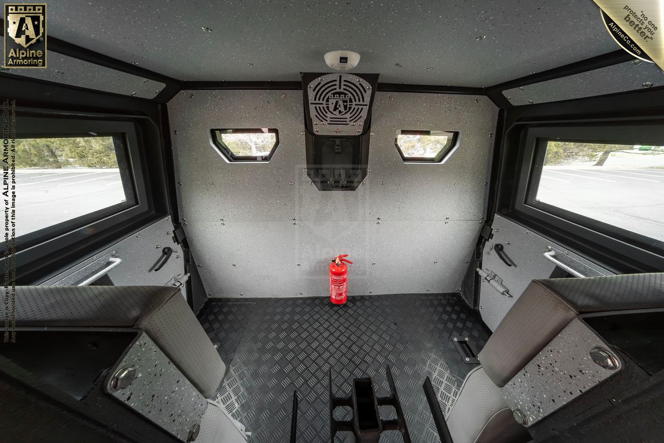 Interior of a vehicle with a fire extinguisher mounted on the wall, two side windows, and two seats on opposite sides. The walls and flooring is made of durable, protective material.