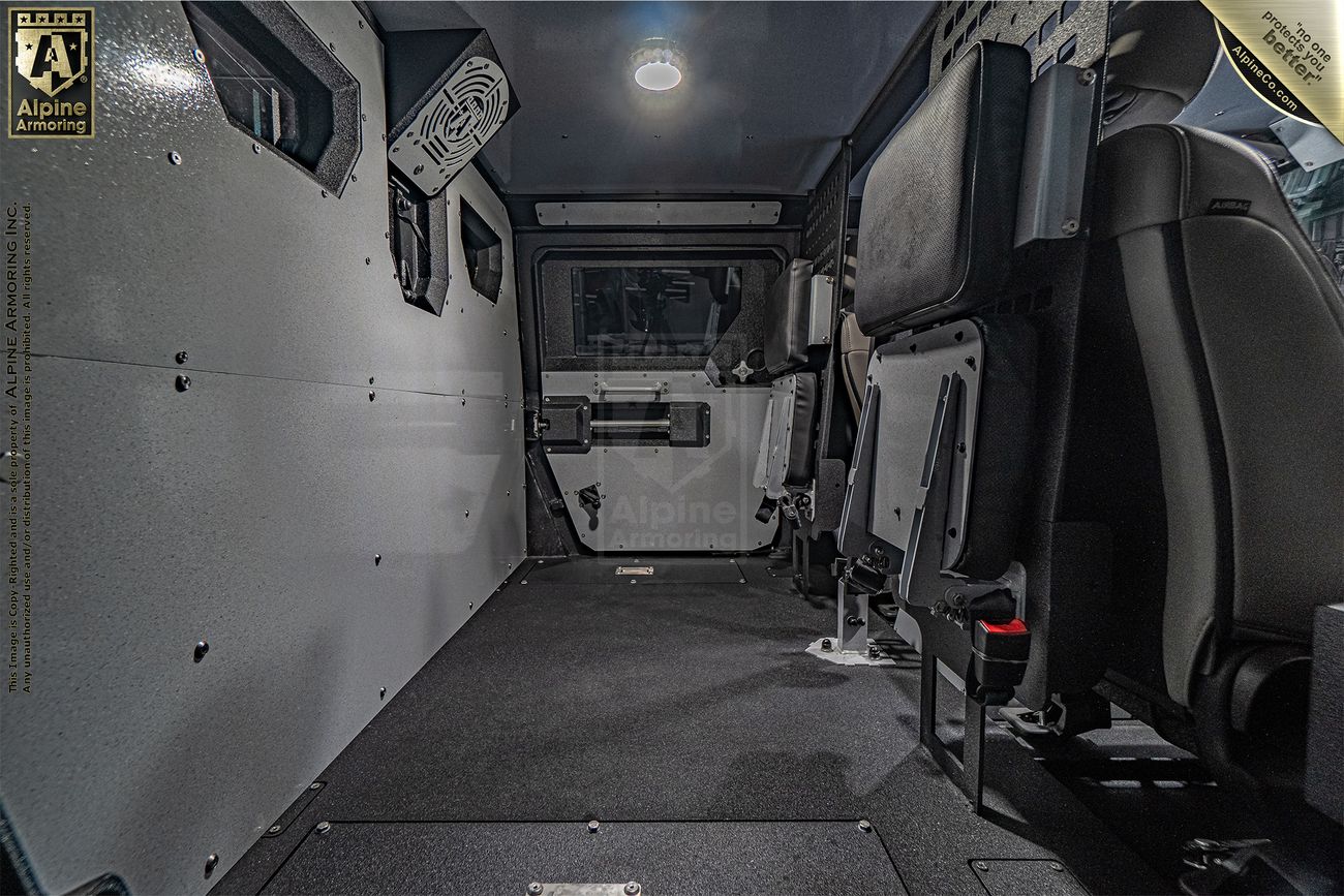 Interior view of an armored Pit-Bull VXT from Alpine Armoring showing a spacious, empty rear cabin with metal walls, a light on the ceiling, and two foldable seats on the right.