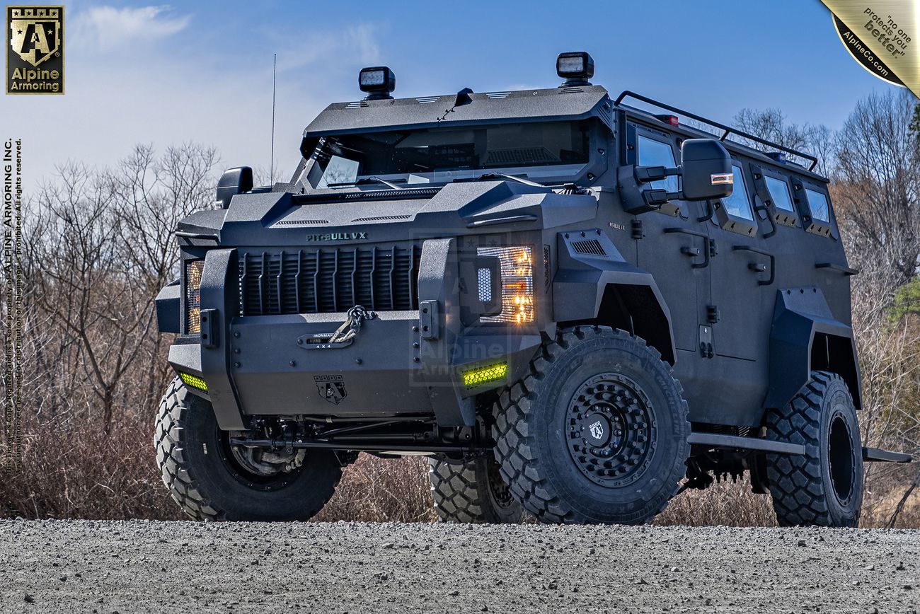 A black, off-road armored Pit-Bull VX APC vehicle with rugged tires is parked on a gravel surface. The vehicle has heavy-duty bumpers, a roof-mounted light bar, and branding that reads "Alpine Armoring.
