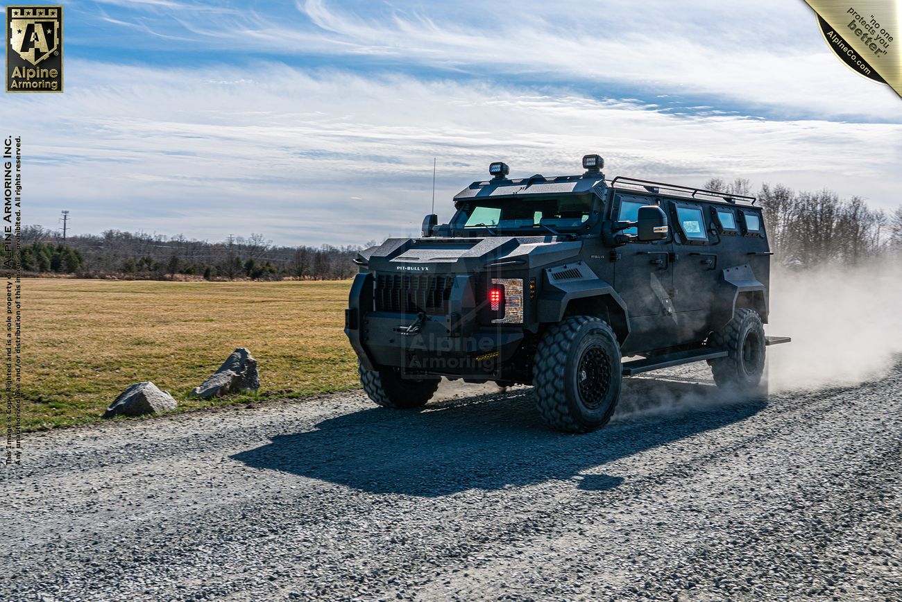 An armored Pit-Bull VX APC drives on a gravel road in a rural area, kicking up dust. The logo of Alpine Armoring is visible on the vehicle and in the top corners of the image.