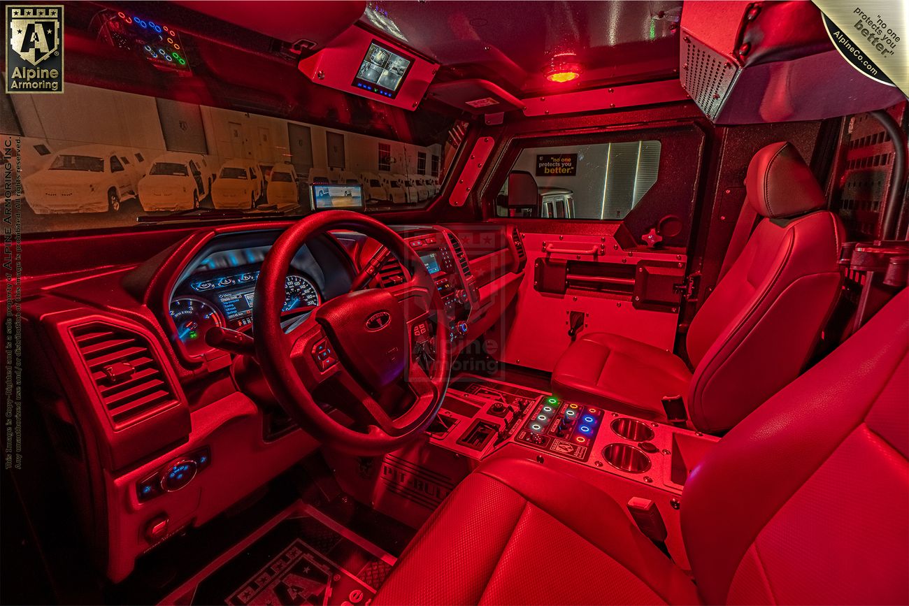 Interior of an armored Pit-Bull VX APC cockpit illuminated by red lighting, showing a steering wheel, dashboard with controls and displays, and two front seats.